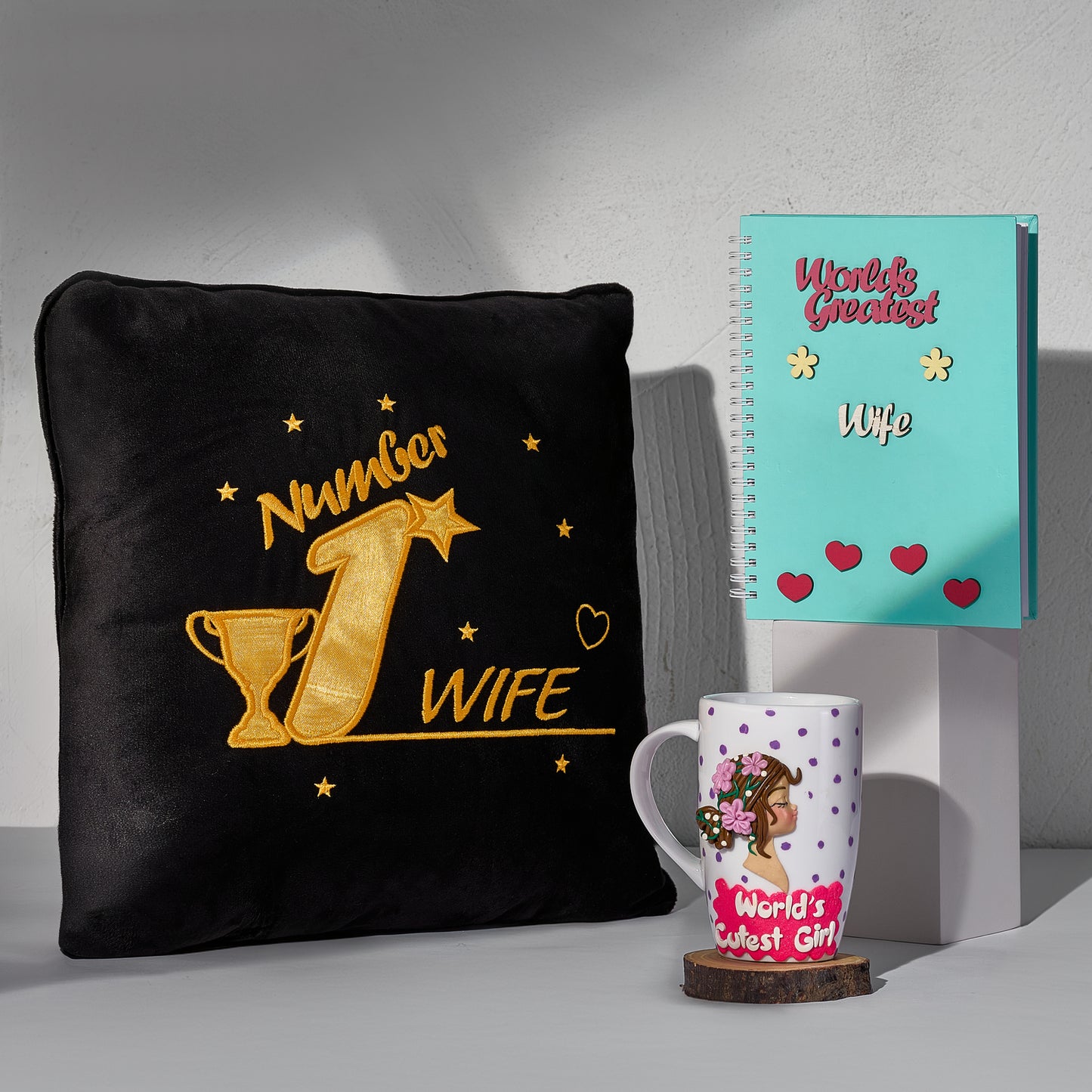 A gift bundle consisting of a black pillow with text 'NUMBER 1 WIFE', a blue notebook titled 'WIFE', and a mug with a graphic of a woman's face and the text 'World's Cutest Girl'. The items are displayed against a light background.